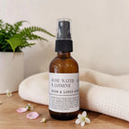 An amber glass room and linen spray bottle with a black spray top and a white label reading 'ROSE WATER & JASMINE ROOM & LINEN SPRAY', styled on a wooden surface with a white textured cloth, a potted fern, and small white flowers.