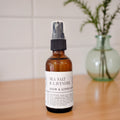 An amber glass room spray bottle labeled 'SEA SALT & LAVENDER ROOM & LINEN SPRAY' stands on a wooden surface with a blurred green vase in the background.