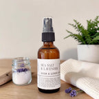 An amber glass room spray bottle labeled 'SEA SALT & LAVENDER ROOM & LINEN SPRAY' stands on a wooden surface, styled with a small jar of lavender bath salts, a folded cream towel, and a potted green plant in the background.