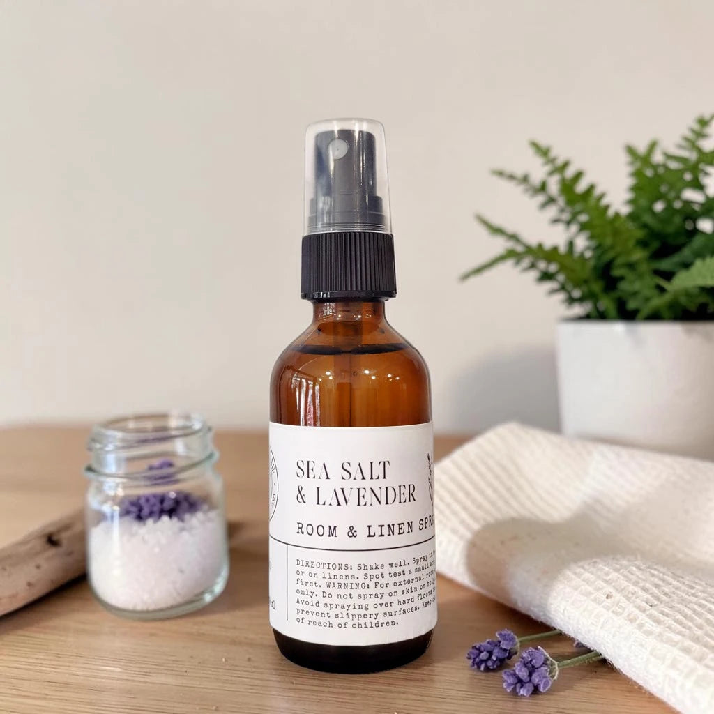 An amber glass room spray bottle labeled 'SEA SALT & LAVENDER ROOM & LINEN SPRAY' stands on a wooden surface, styled with a small jar of lavender bath salts, a folded cream towel, and a potted green plant in the background.