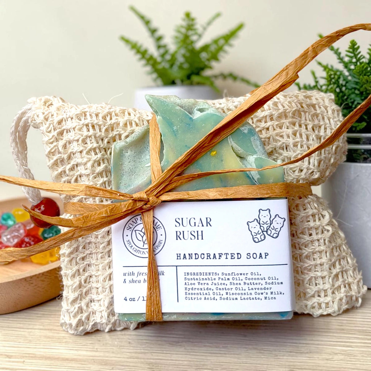 Sugar Rush Essentials Gift Set: a blue swirled soap bar tied with raffia in a natural sisal bag, surrounded by colorful candy pieces on a wood surface.