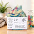 A handmade sugar rush soap bar with colorful swirls of pink, blue, green, and yellow, resting on a wooden surface with its white paper label. The background features a soft-focus plant and a glass jar of colorful gummy bears.