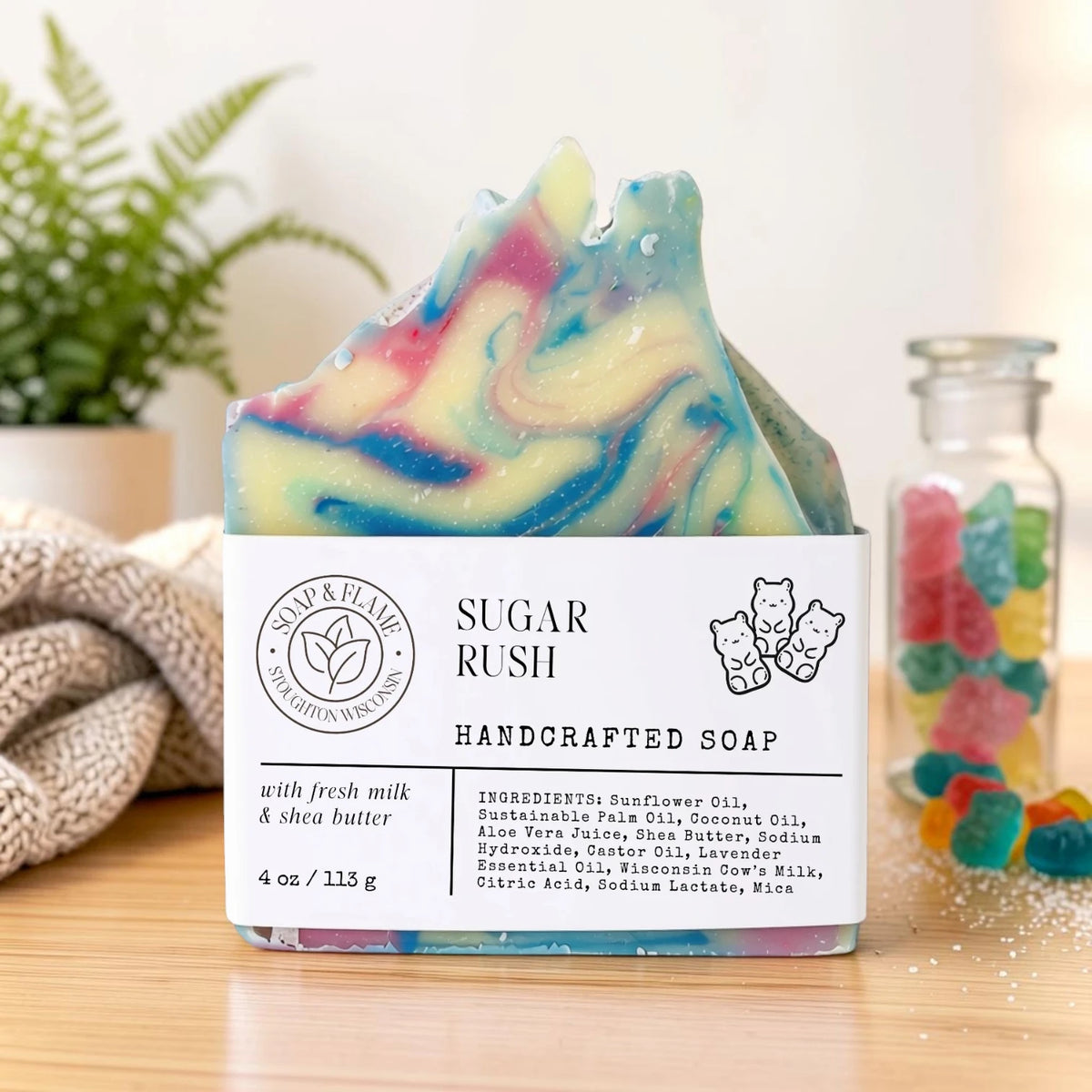A handmade sugar rush soap bar with colorful swirls of pink, blue, green, and yellow, resting on a wooden surface with its white paper label. The background features a soft-focus plant and a glass jar of colorful gummy bears.
