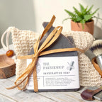 The Barbershop Essentials Gift Set: a charcoal soap bar tied with raffia in a natural sisal bag, styled with a wooden shaving brush and a small wood bowl against a dark background.