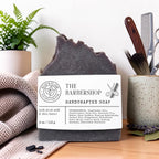 the barbershop soap on a wood table with a knit blanket, fern and a ceramic mug with barber items in it.
