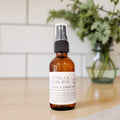 An amber glass spray bottle labeled 'Vanilla & Dark Rum Room & Linen Spray' with a black spray top, standing on a light wood surface with a blurred white tile wall and green plant in the background.