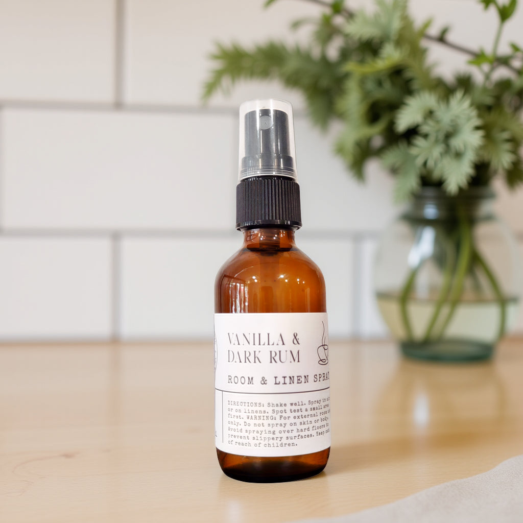 An amber glass spray bottle labeled 'Vanilla & Dark Rum Room & Linen Spray' with a black spray top, standing on a light wood surface with a blurred white tile wall and green plant in the background.