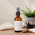An amber glass room and linen spray bottle with a white label reading 'Vanilla & Dark Rum' and 'Room & Linen Spray,' styled on a tabletop with cinnamon sticks, a folded towel, small glass jars, and a plant in the background.