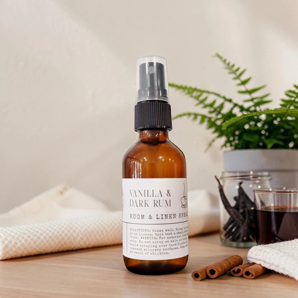 An amber glass room and linen spray bottle with a white label reading 'Vanilla & Dark Rum' and 'Room & Linen Spray,' styled on a tabletop with cinnamon sticks, a folded towel, small glass jars, and a plant in the background.
