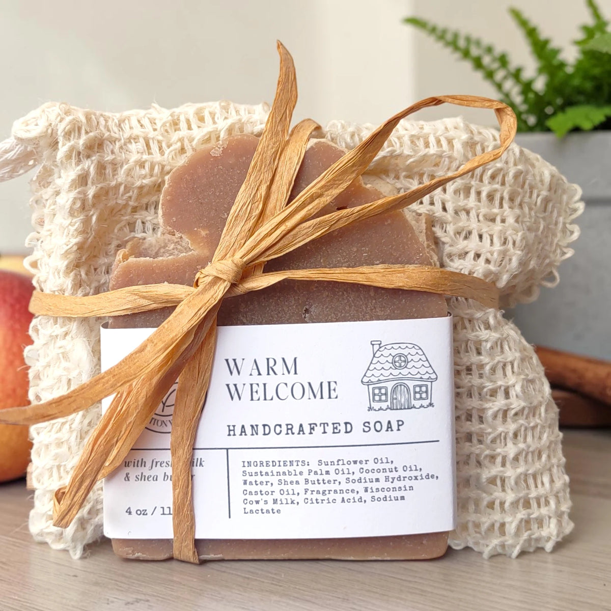 Warm Welcome Essentials Gift Set: a soap bar tied with raffia in a natural sisal bag, styled with a small apple and greenery on a wood surface.