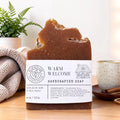 A handcrafted bar of soap with a rustic broken top, labeled 'Warm Welcome Handcrafted Soap'. The soap is brown and sits on a wooden surface with a cozy background featuring a knit blanket, a potted fern, and a ceramic mug.