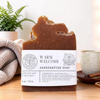 A handcrafted bar of soap with a rustic broken top, labeled 'Warm Welcome Handcrafted Soap'. The soap is brown and sits on a wooden surface with a cozy background featuring a knit blanket, a potted fern, and a ceramic mug.