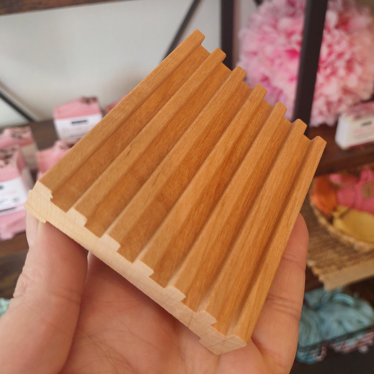 A hand holding a rectangular wooden soap dish with a ribbed, slatted top surface, shown against a blurred indoor background.